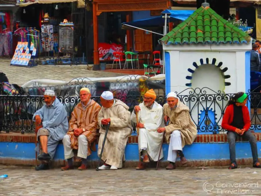 5 men in djellabas sitting on a blue wall in a town square