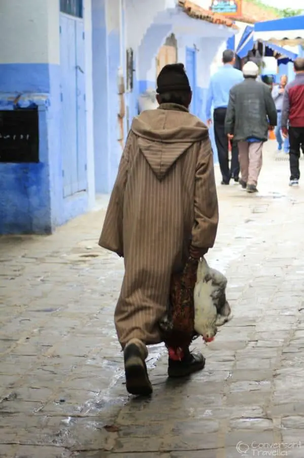 Man in a cloak walking down a street carrying a chicken