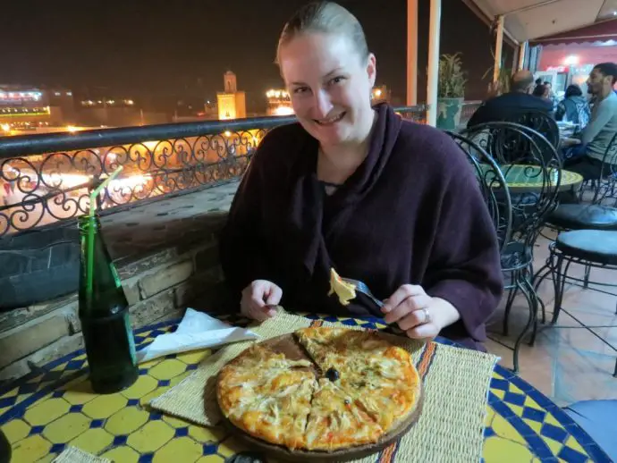 Woman eating pizza at a table on the balcony of a restaurant overlooking a night city skyline