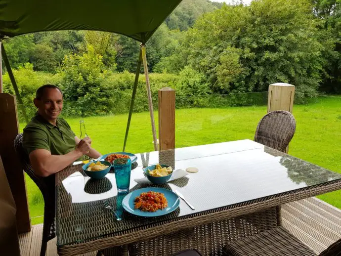 Man sitting at a table eating dinner on the wooden deck of a glamping tent with views of grassy field and trees - glamping in Wales