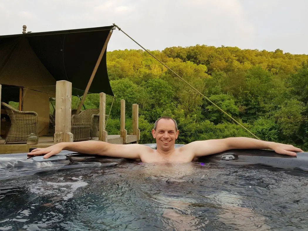 Smiling man in hot tub with glamping tent and forest in the background