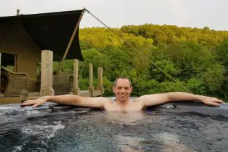 Smiling man in hot tub with glamping tent and forest in the background