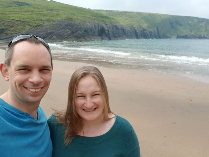 Two people smiling on a beach with cliffs in the background and sea to the right - glamping in wales