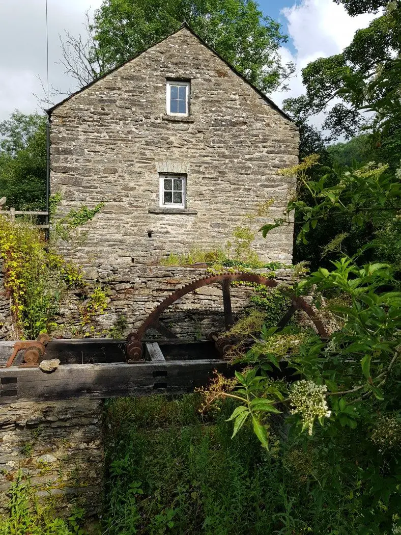 Old stone mill building with 2 small windows at the top, and a large rusty water wheel at the bottom