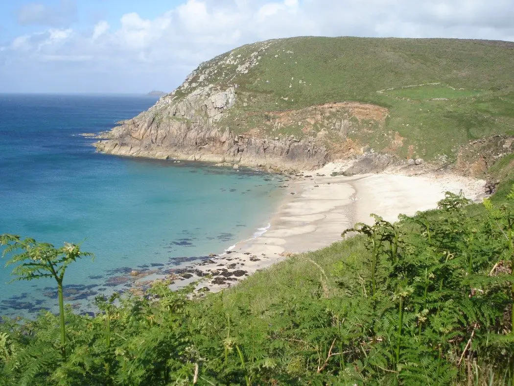 Moon shaped beach with rocky headland in the background, grass in the foreground and sea to the left - Portheras Cove in Cornwall