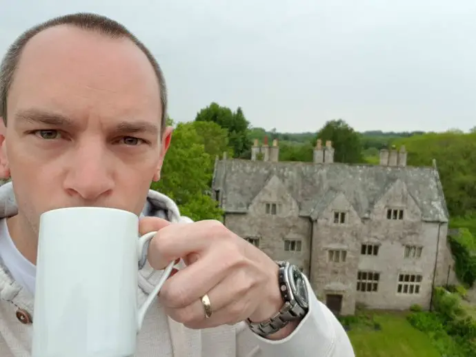 Man standing on top of a gatehouse tower drinking coffee with manor house in the background