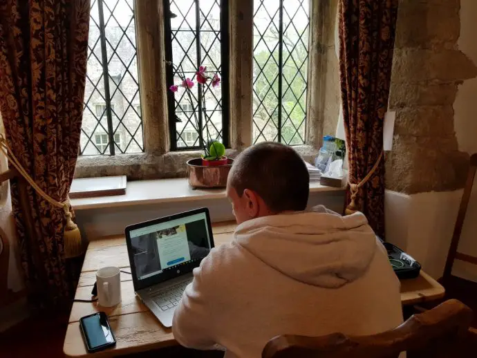 Man in white jumper sitting at table looking at laptop in front of big window