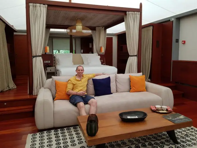 Room interior with 4 poster bed and man in yellow shirt sitting on a sofa in front of it