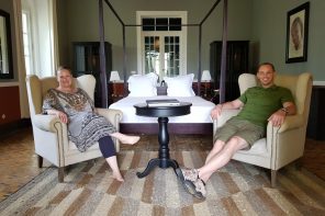 Bedroom with 4 poster bed, and 2 people sitting in armchairs in front of it