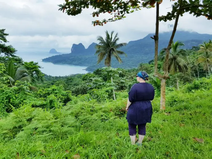 Woman standing facing the ocean surrounded by tropical green vegetation and palm trees