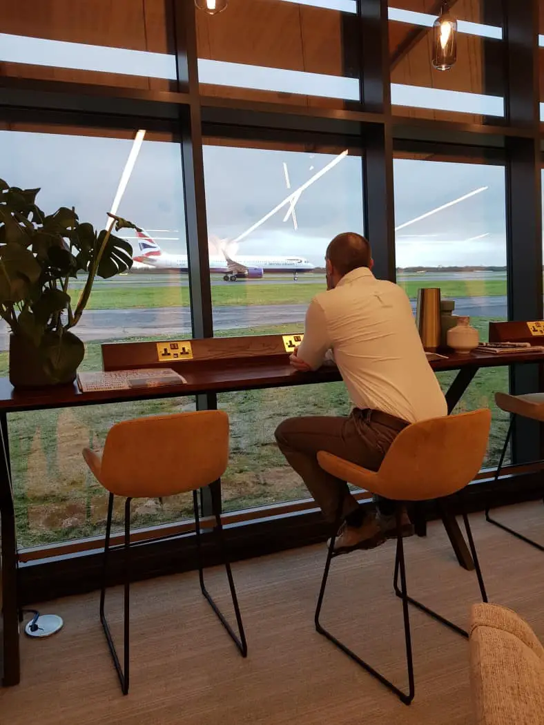 Man sitting on a high stool looking out of large window as a plane passes on the runway