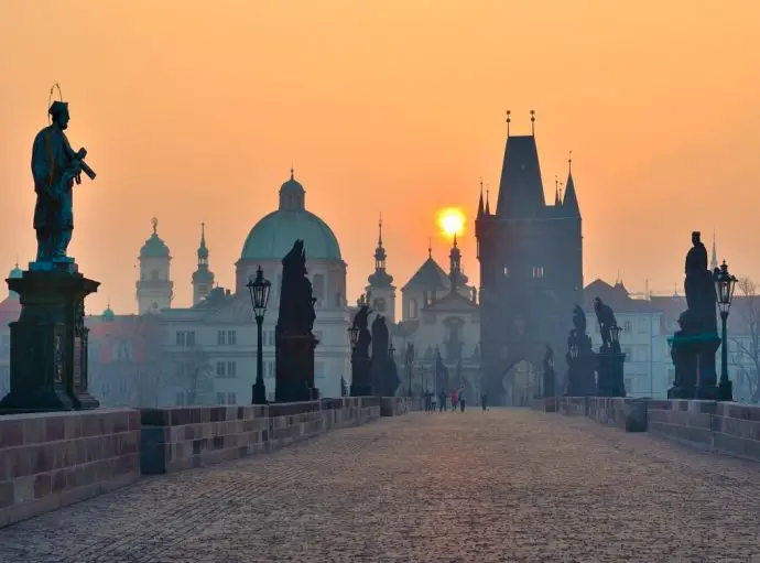 Bridge with stone statues along the edges leading towards a tower at sunset