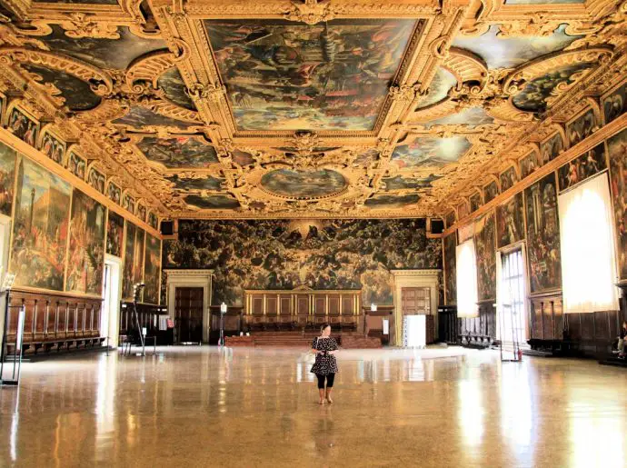 Woman standing alone in the middle of a large museum hall with ornate gold ceilings and murals. 