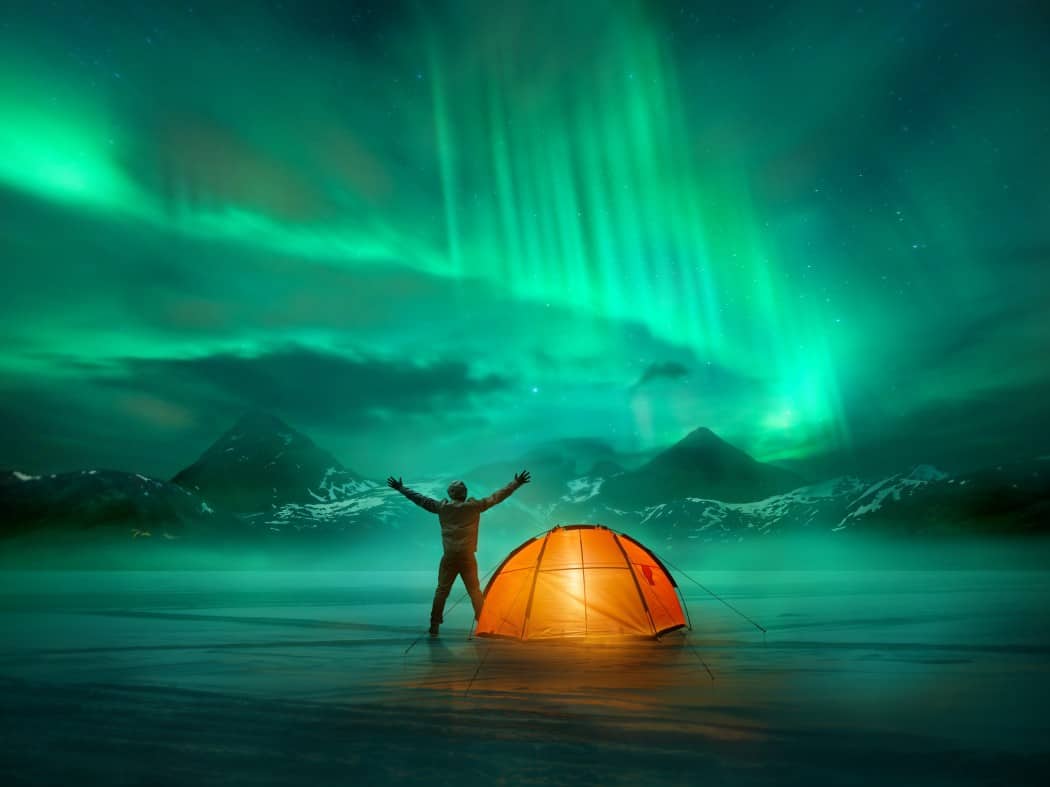 Man standing with out-stretched arms beside an orange tent looking towards northern lights over the mountains