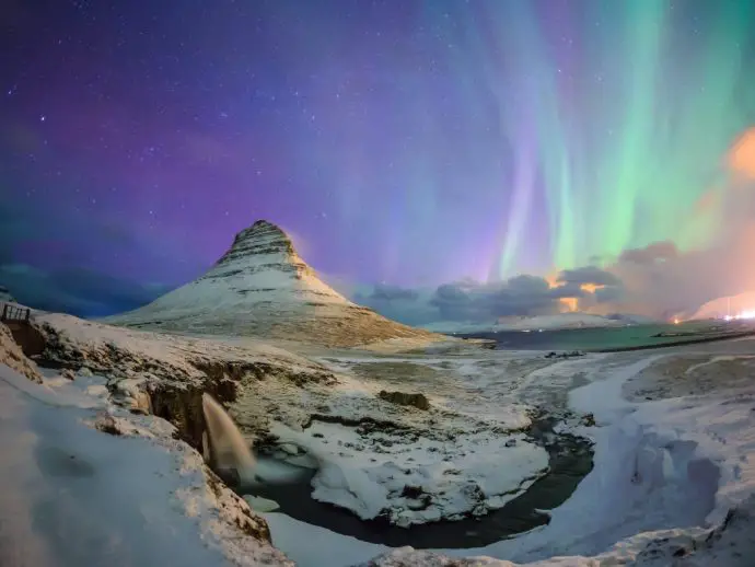 Snowy mountain peak with a river in the foreground and northern lights in the sky in the background