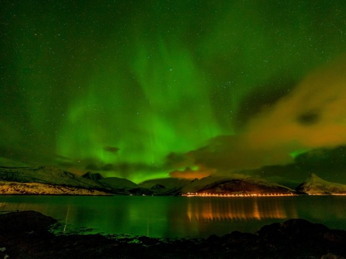 Green northern lights in the sky above a town with lit up buildings against a dark mountain backdrop