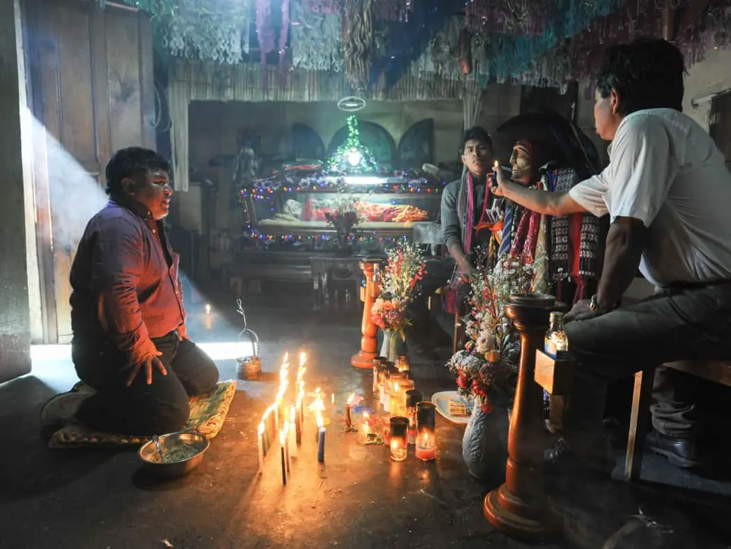 Small group of men in a darkened room, one on the left is kneeling to pray to a wooden effigy of a colourfully dressed saint