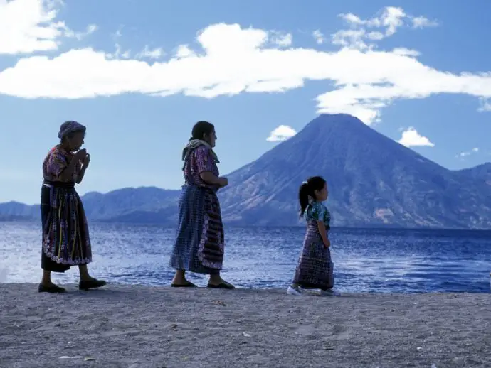 Two women and a small girl walking in line along a sandy beach in front of a lake with a volcano in the background