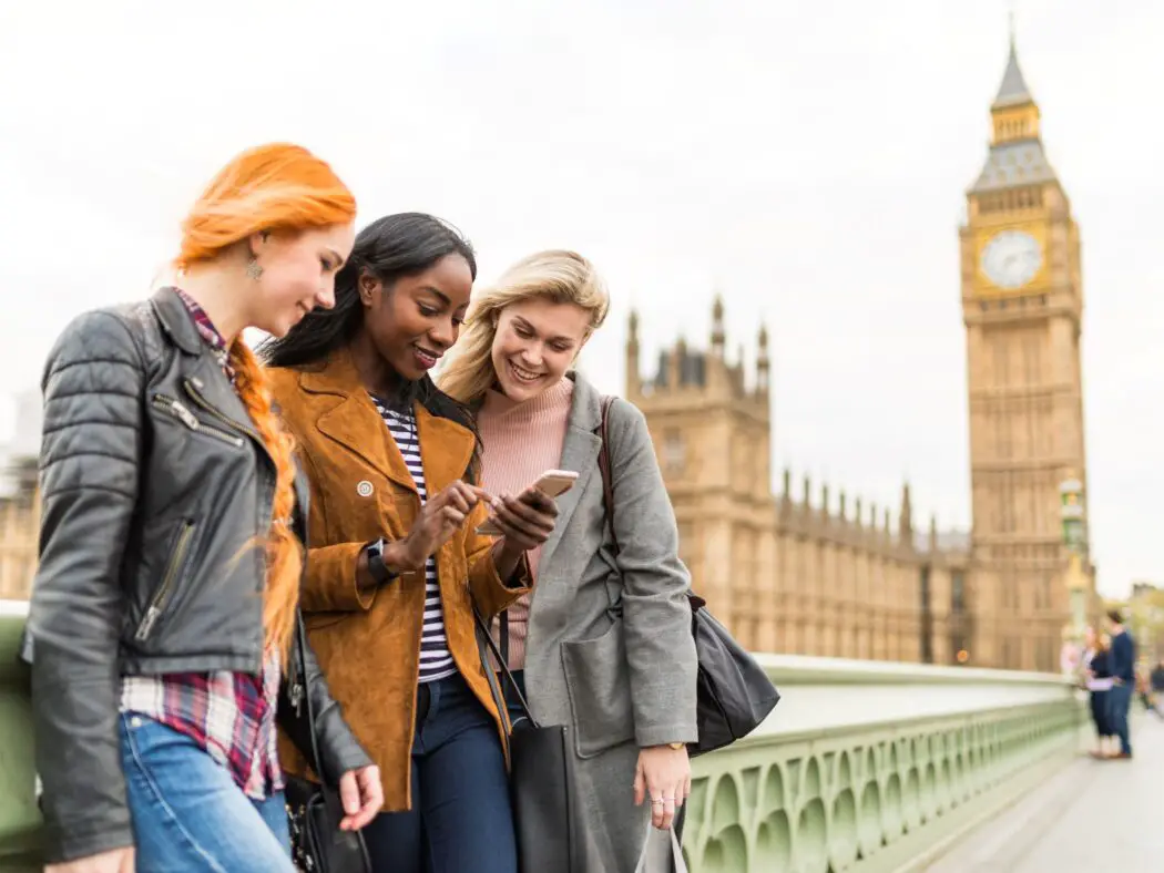 Three ladies looking at their phones with a stone tower with a clock at the top in the background