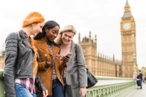 Three ladies looking at their phones with a stone tower with a clock at the top in the background